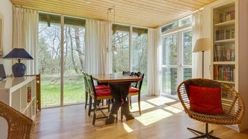 Une pièce spacieuse avec un plancher en bois, une table à manger avec des chaises, un fauteuil en osier avec un coussin rouge, une bibliothèque et de grandes fenêtres donnant sur l’extérieur.