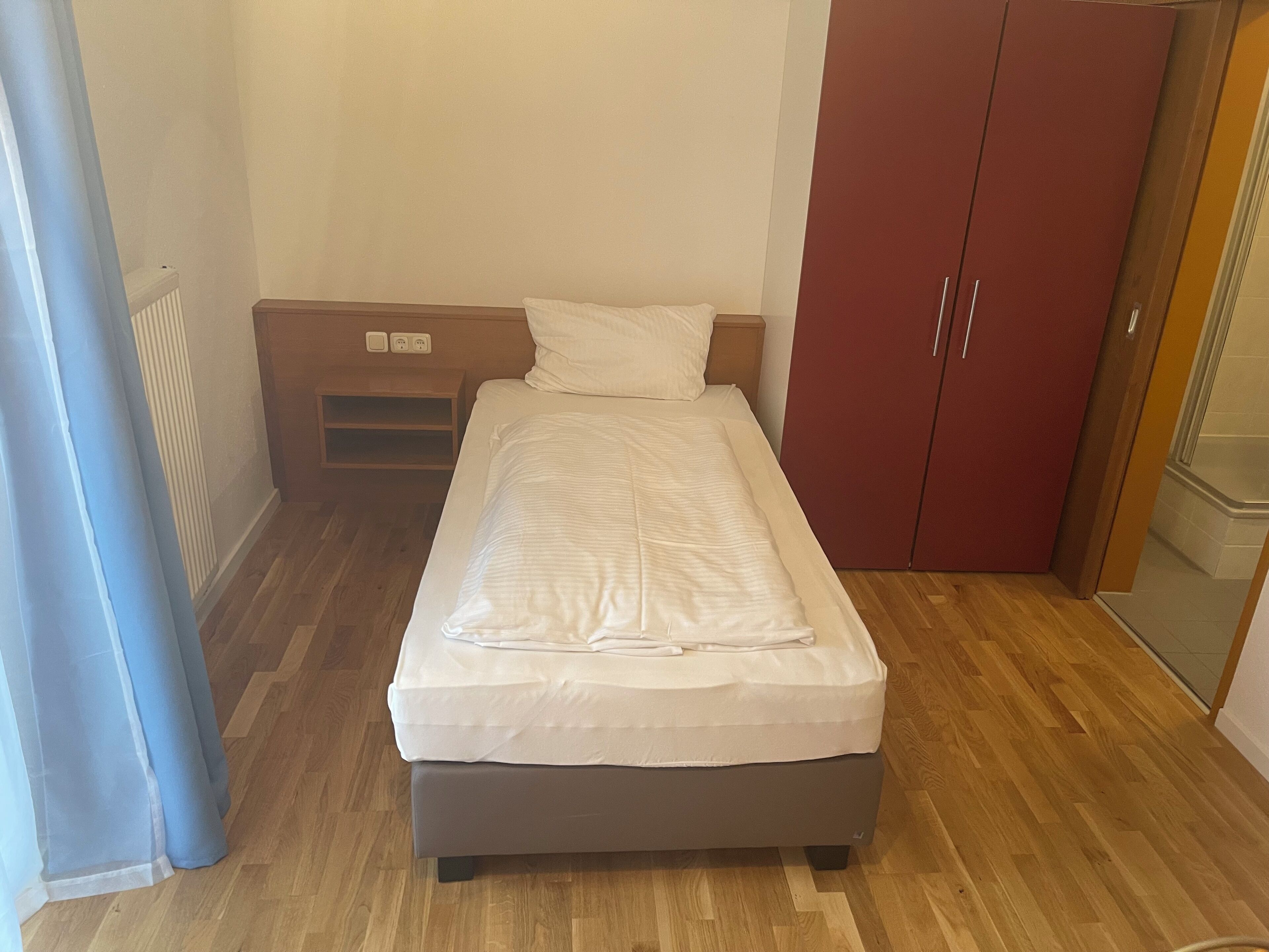 Un petit lit bien fait dans une chambre compacte, avec un plancher en bois, une armoire rouge et une petite table de chevet équipée d’une prise électrique.