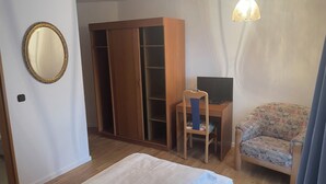 A bedroom with a bed, a wooden wardrobe, a desk with a chair, a television, and a patterned armchair. - Furnished apartment for 1 person (Büchlberg)
