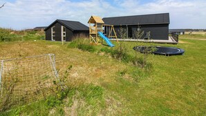 Ein modernes Gebäude mit schwarzem Äußeren, einer hölzernen Spielanlage mit Rutsche und einem Trampolin auf einer Rasenfläche.