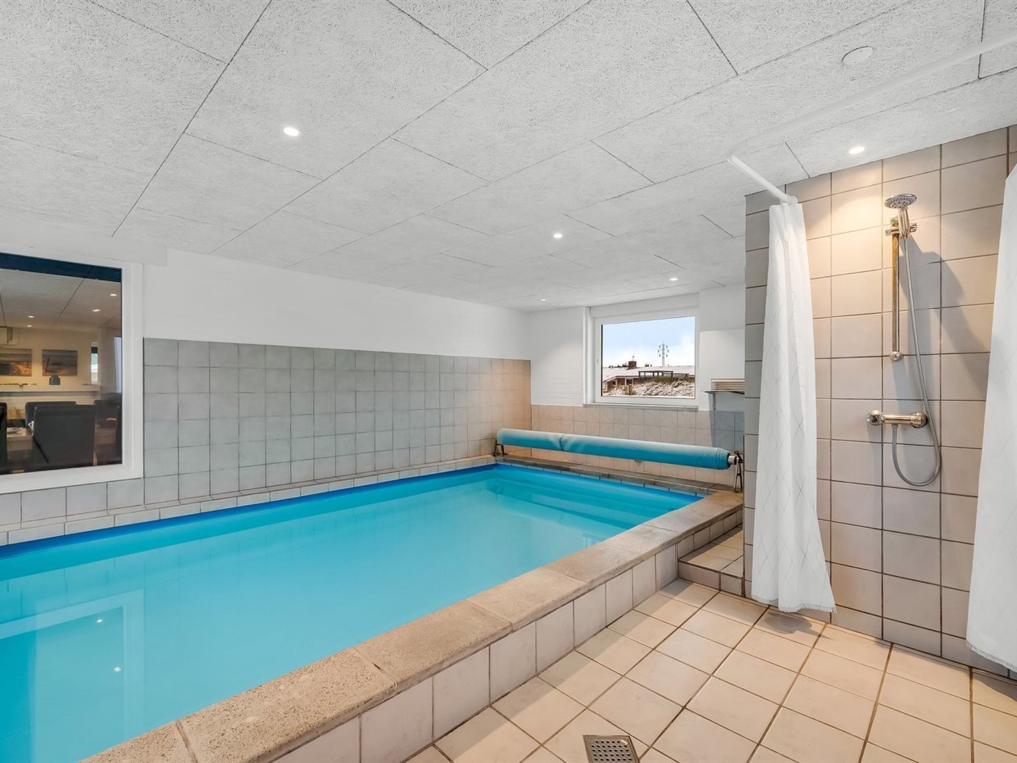 A swimming pool with a shower area, tiled walls, and a window with a view.