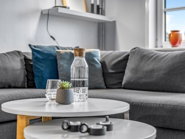 A modern living room with a grey sofa, a round white coffee table, a clear glass bottle, a small potted plant, and a few glasses.