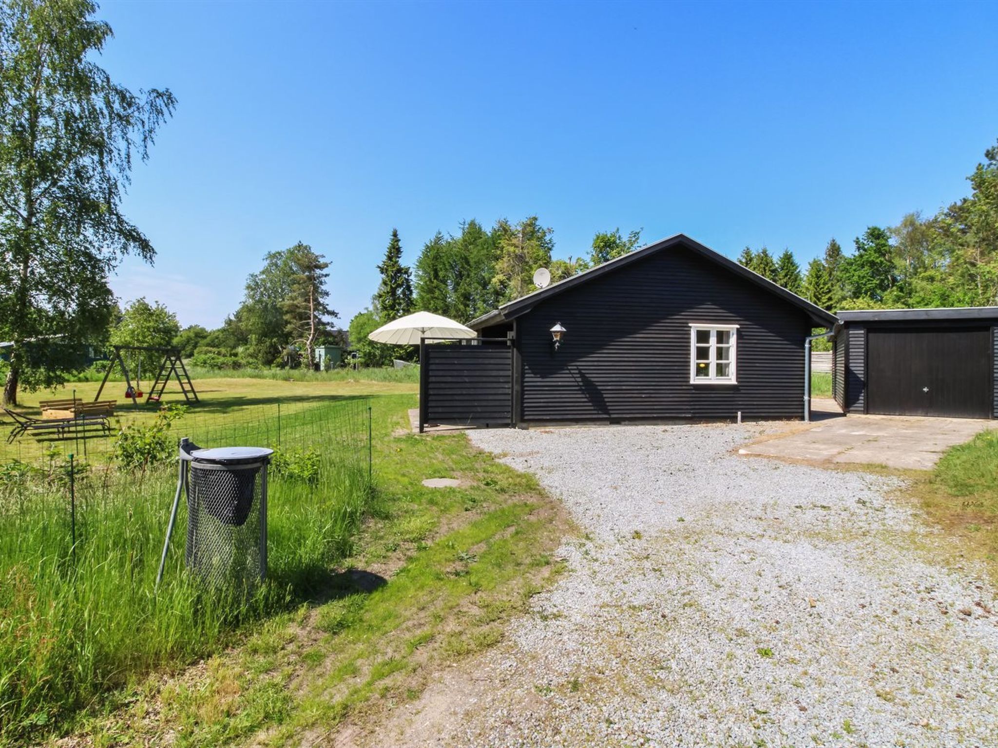 Een zwarte houten chalet met een grindpad, een schommel en een omheind terrein.