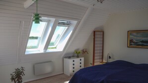 A bedroom with a sloped ceiling, skylights, a bed with a blue cover, a white dresser, a small table with a plant, and a painting on the wall. - "Harre" - 300m from the sea by Interhome (Rudkøbing)