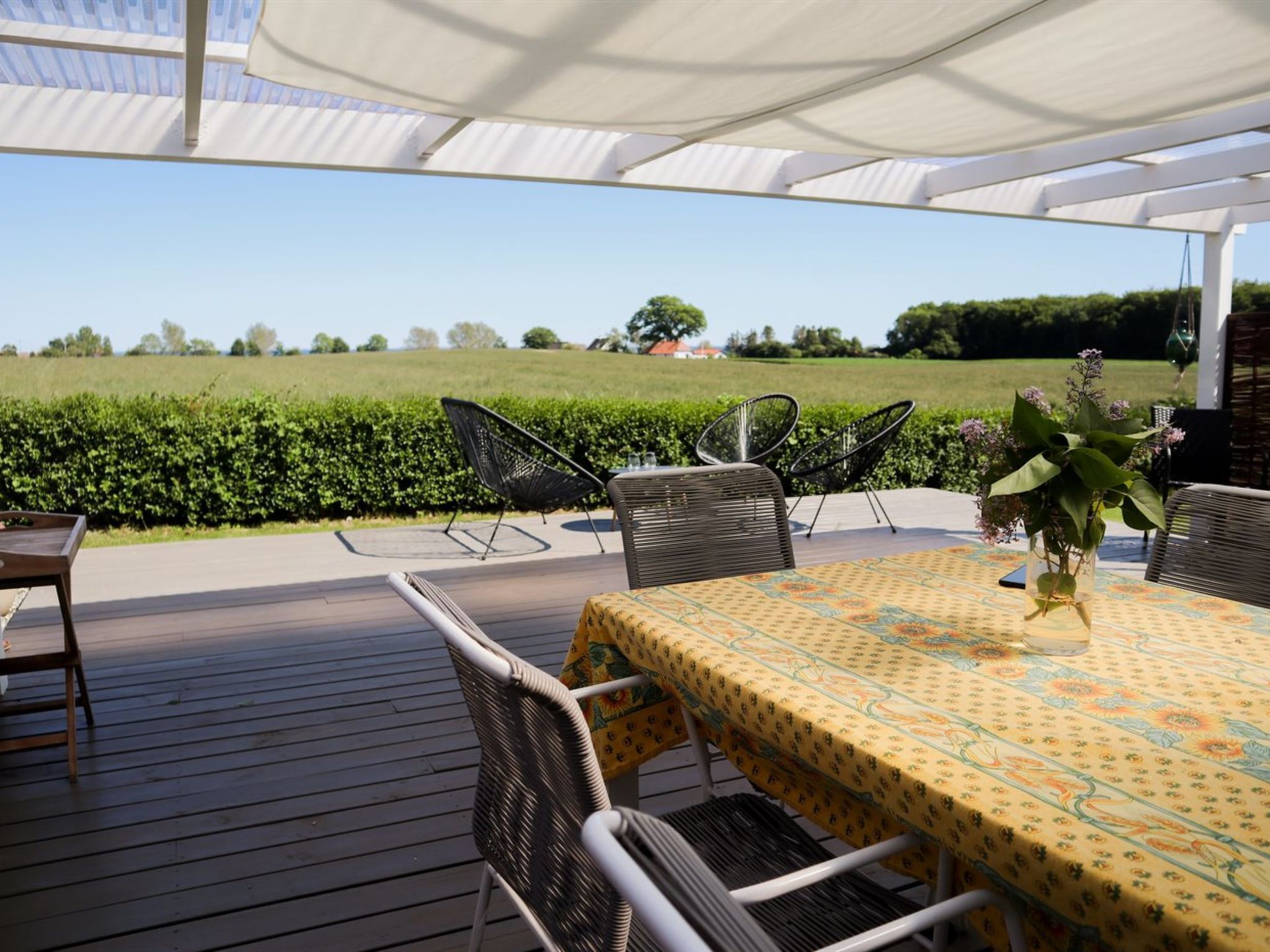 En udendørs terrasse med et bord og stole, et overdækket område og udsigt til en grøn mark.