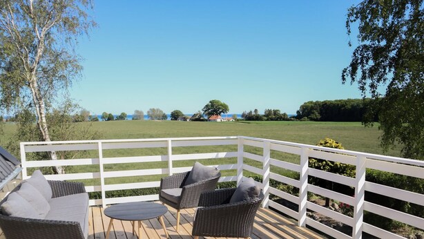 A wooden deck with wicker furniture and a white railing, overlooking a green landscape and a body of water. - "Harre" - 300m from the sea by Interhome (Rudkøbing)