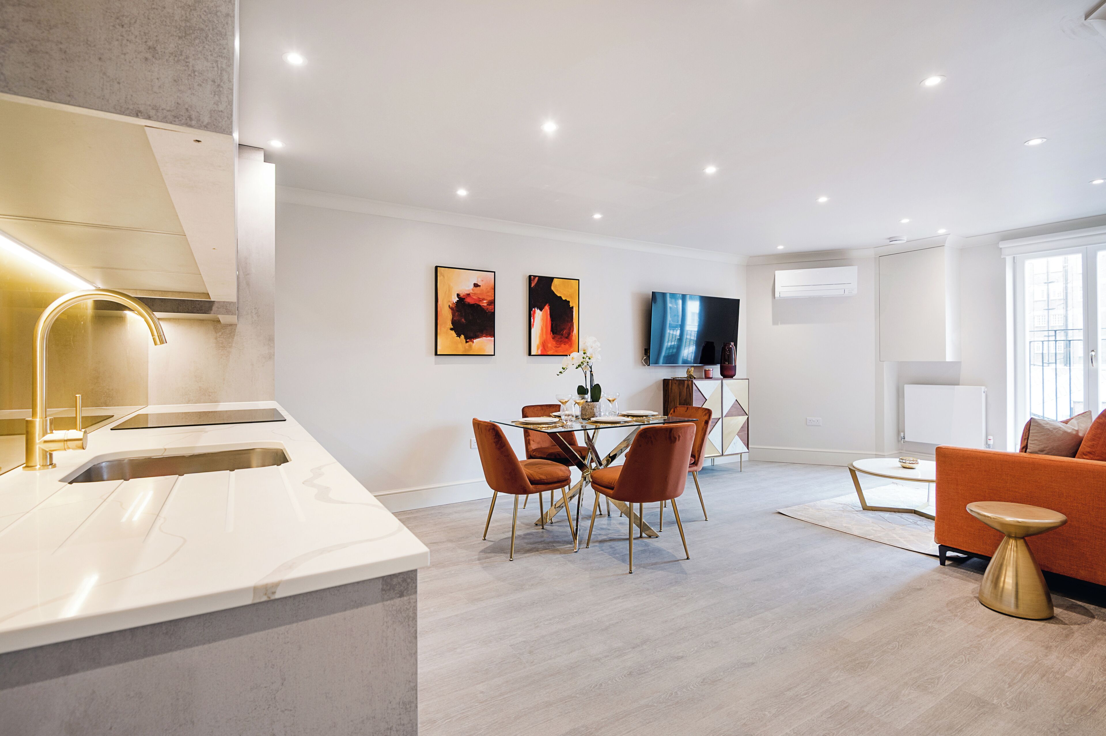 Une cuisine moderne dotée d’un évier encastré, un coin repas avec une table en verre et des chaises orange, un téléviseur à écran plat et un canapé avec des coussins orange.