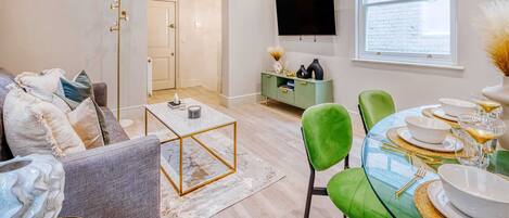 A modern living room with a gray sofa, a glass coffee table, and a green chair. A dining area is visible with a glass table set for four.