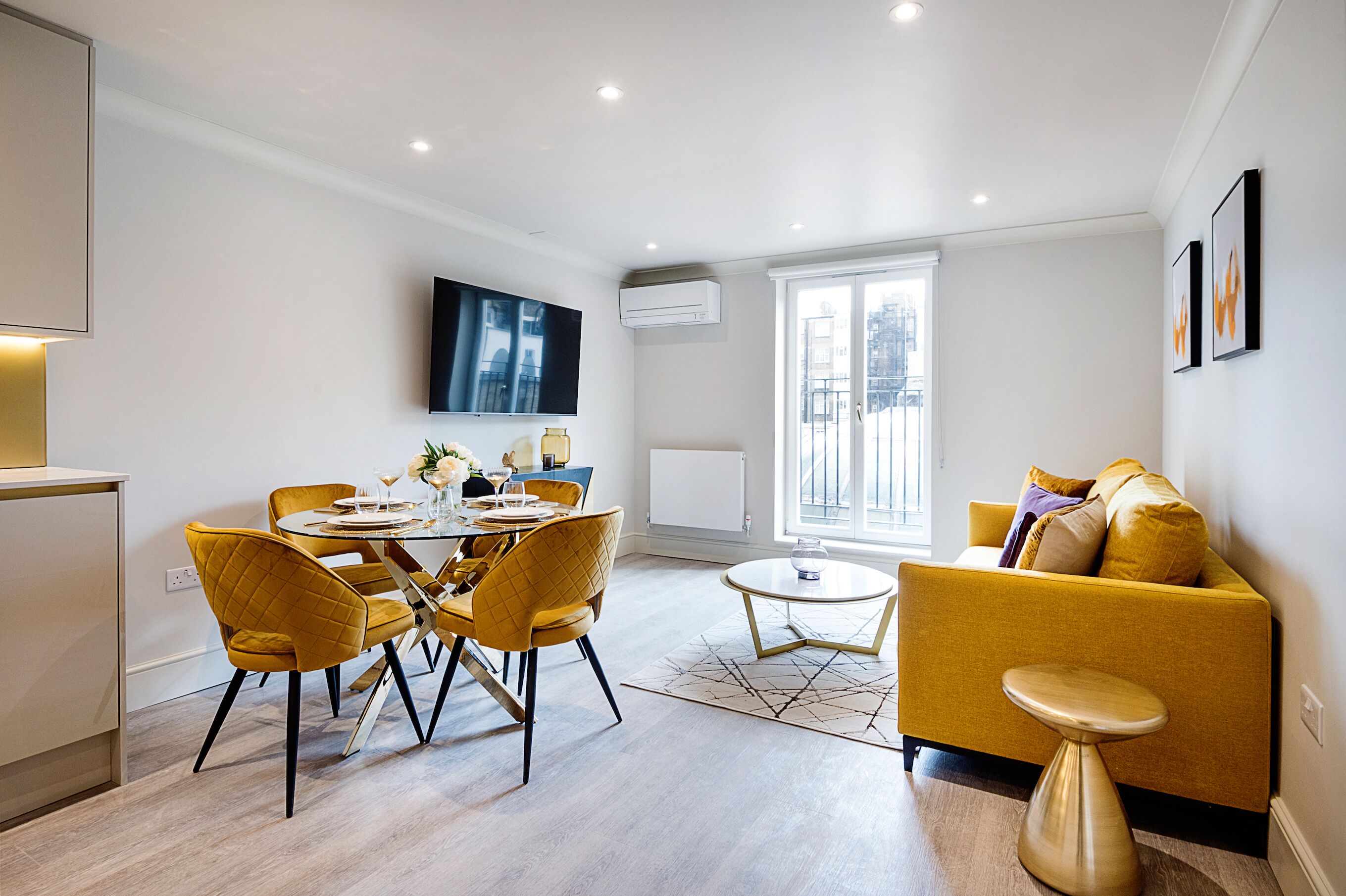 A modern living room with a yellow sofa, a dining table with chairs, and a TV mounted on the wall.