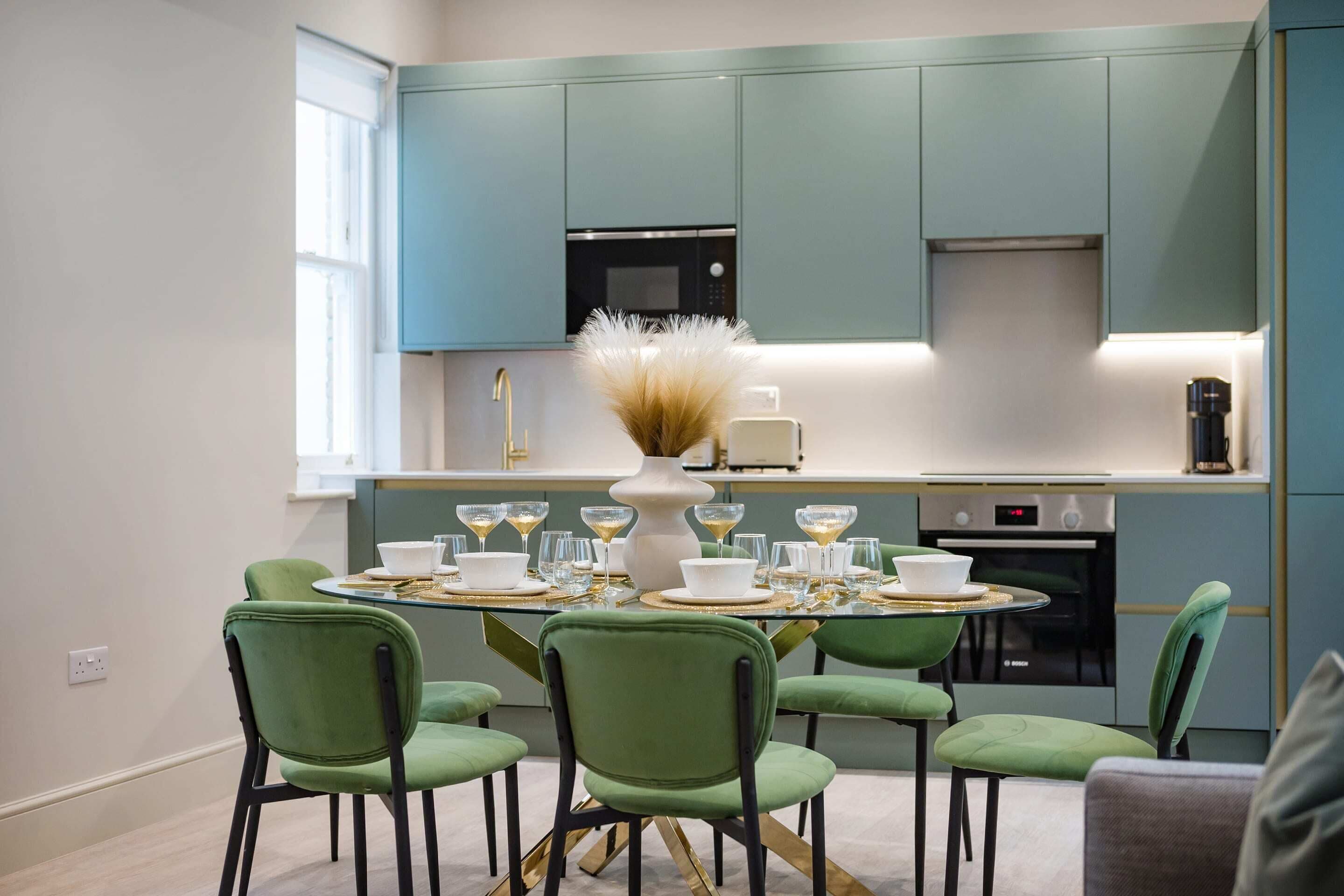 A modern kitchen with teal cabinets, a central island with a vase, and a dining area with green chairs and a glass table.
