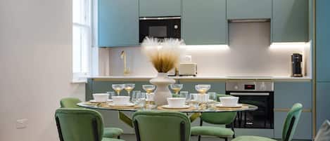 A modern kitchen with teal cabinets, a central island with a vase, and a dining area with green chairs and a glass table.