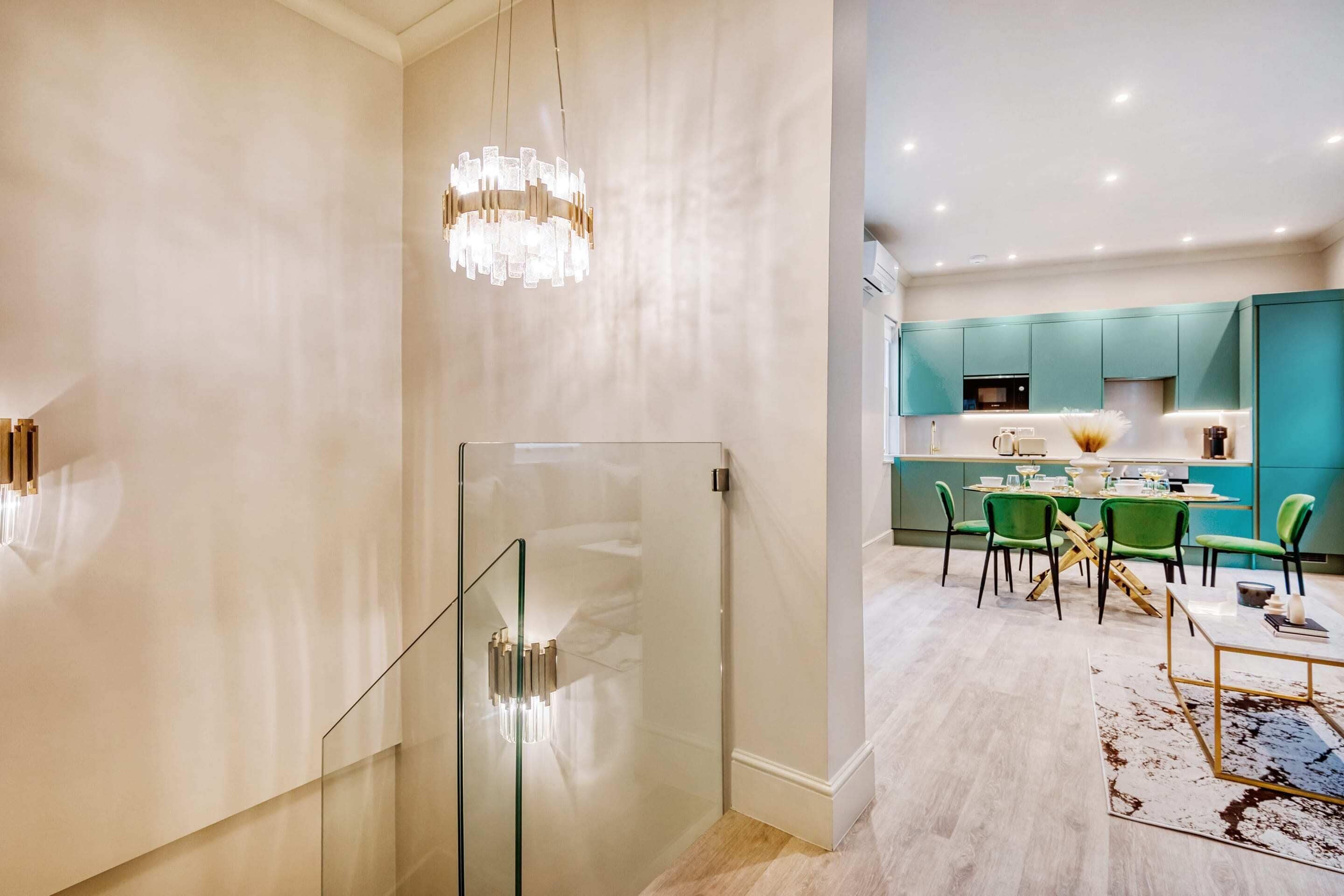 Una cocina moderna con armarios verde azulado, un comedor con sillas verdes y una escalera de cristal.