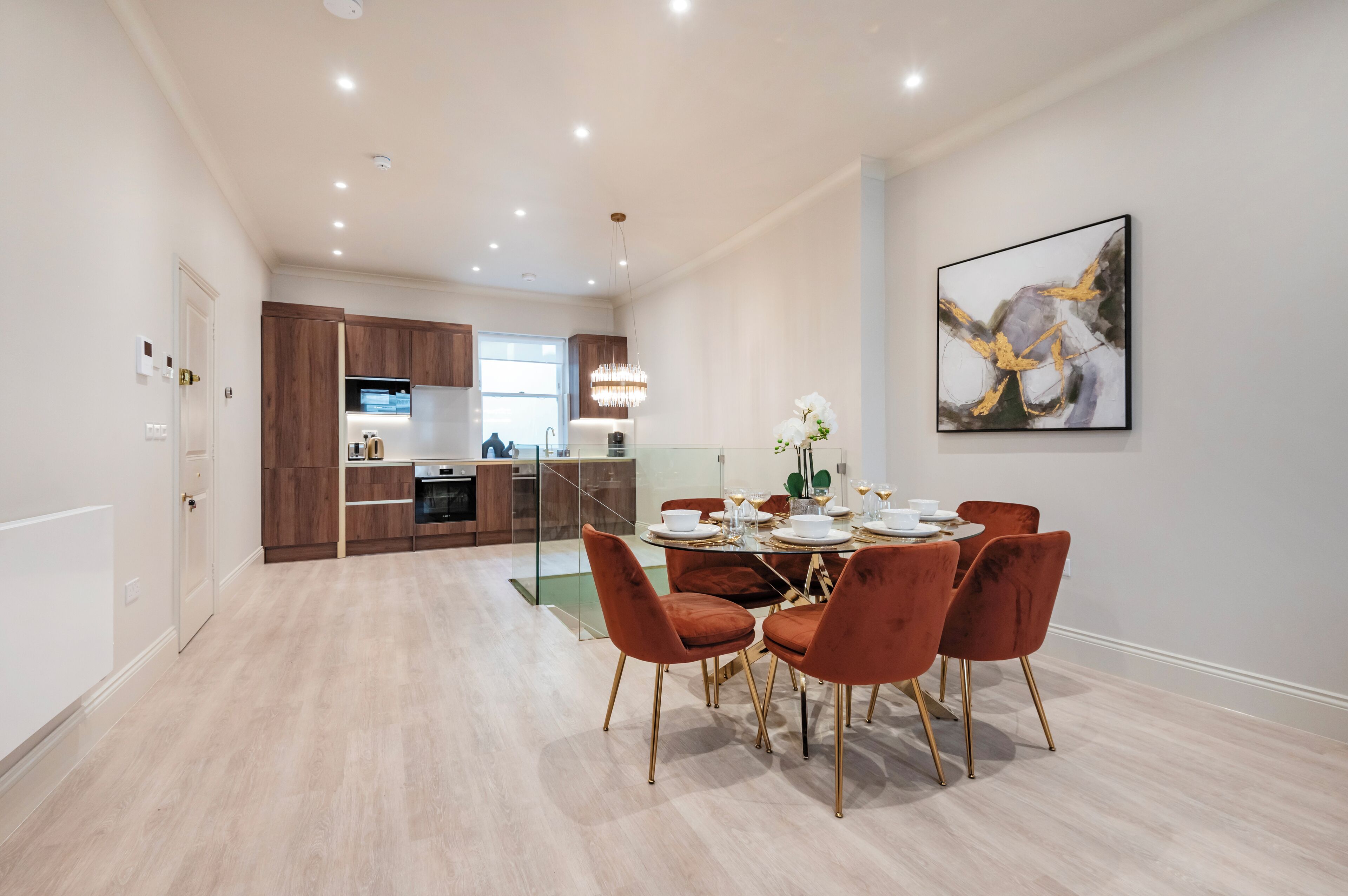 Eine moderne Küche mit Holzschränken, ein Essbereich mit Glastisch und roten Stühlen sowie ein zeitgenössisches Kunstwerk an der Wand.