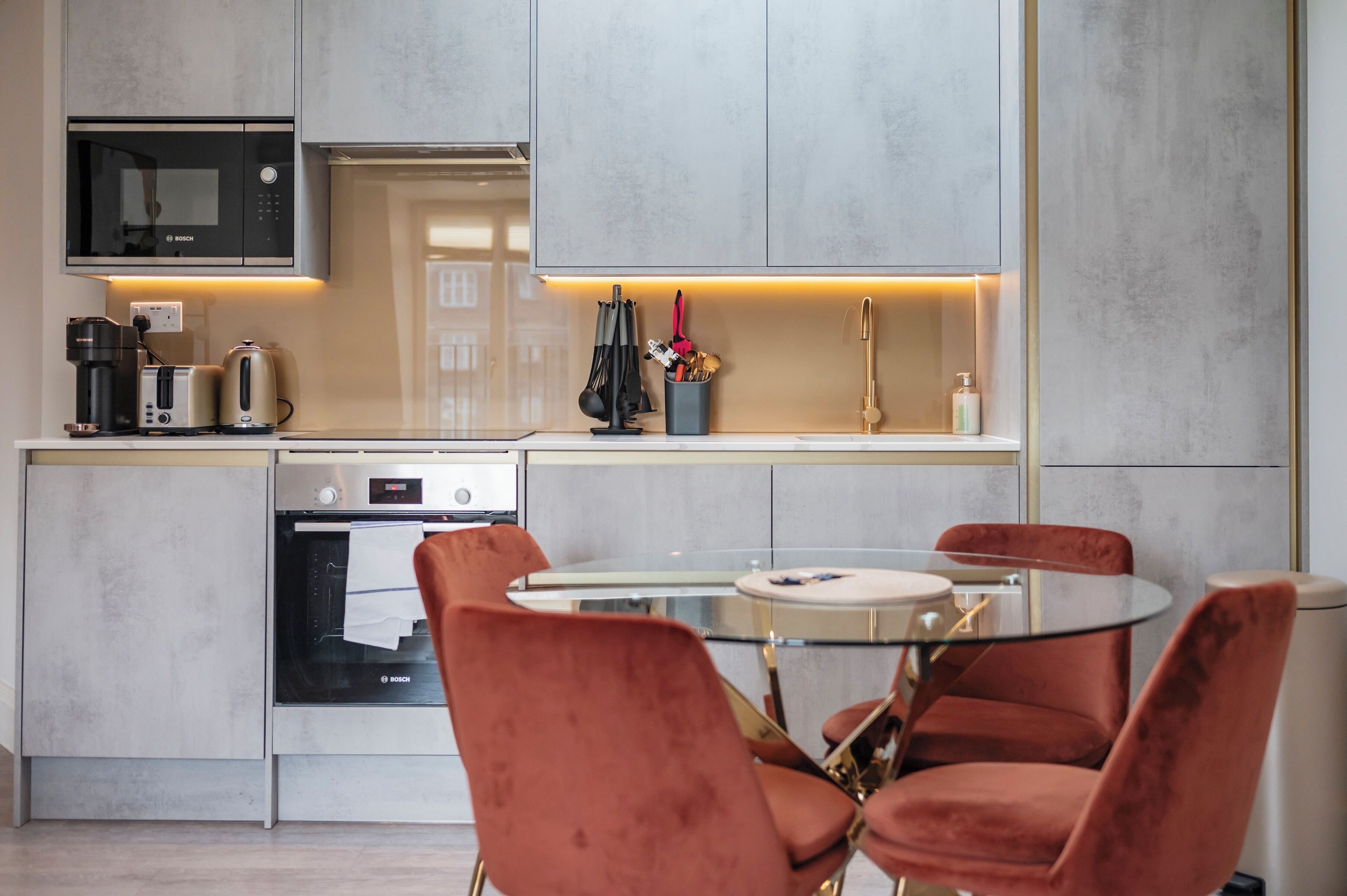 A modern kitchen with a Bosch microwave, oven, and coffee maker, a glass-top dining table with red chairs, and a hanging pot rack.