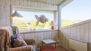 Une pièce chaleureuse avec un plafond en bois, une grande fenêtre donnant sur des maisons, un canapé à motifs, un pouf rouge et une petite table avec une lampe.