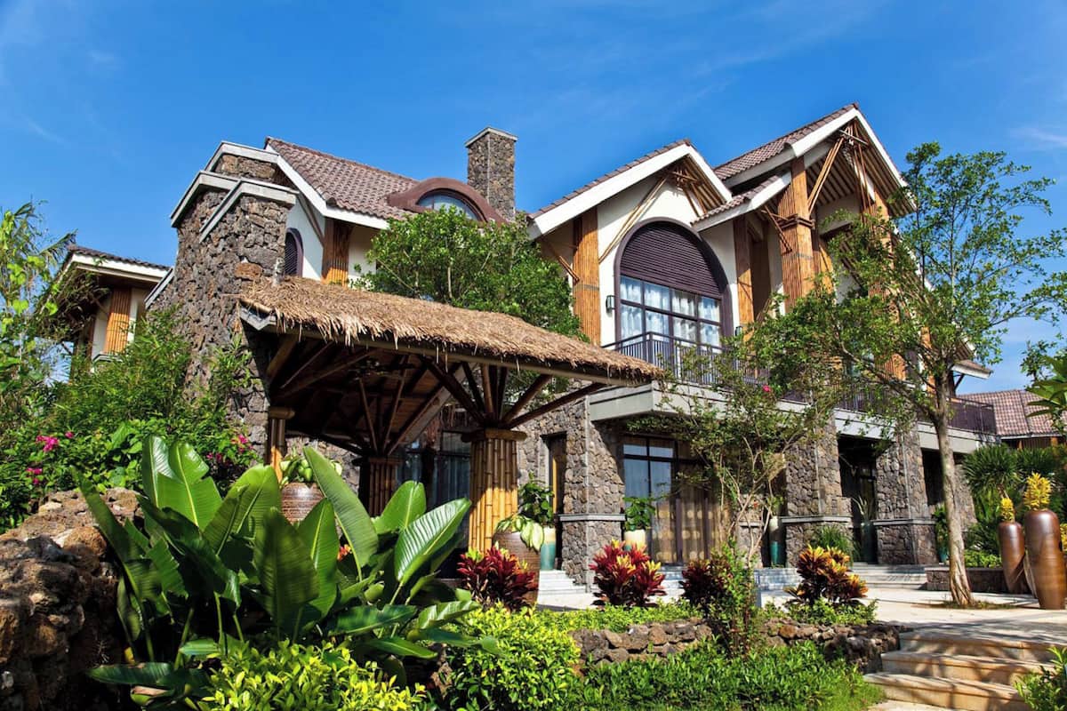 A large, two-story house with a stone facade, a thatched-roof structure, and a well-maintained garden with tropical plants.