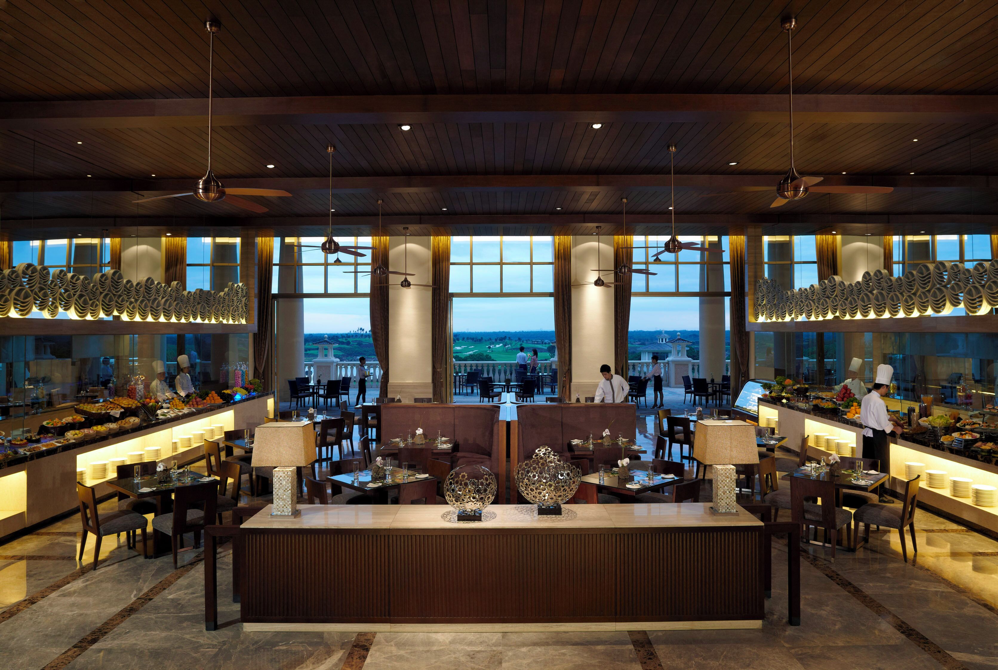A spacious hotel lobby with a central reception desk, multiple dining areas, and large windows offering a view of the cityscape.