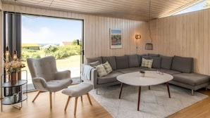 Ein modernes Wohnzimmer mit Sofa, Sessel und rundem Couchtisch, großen Fenstern und einer Holzbalkendecke.