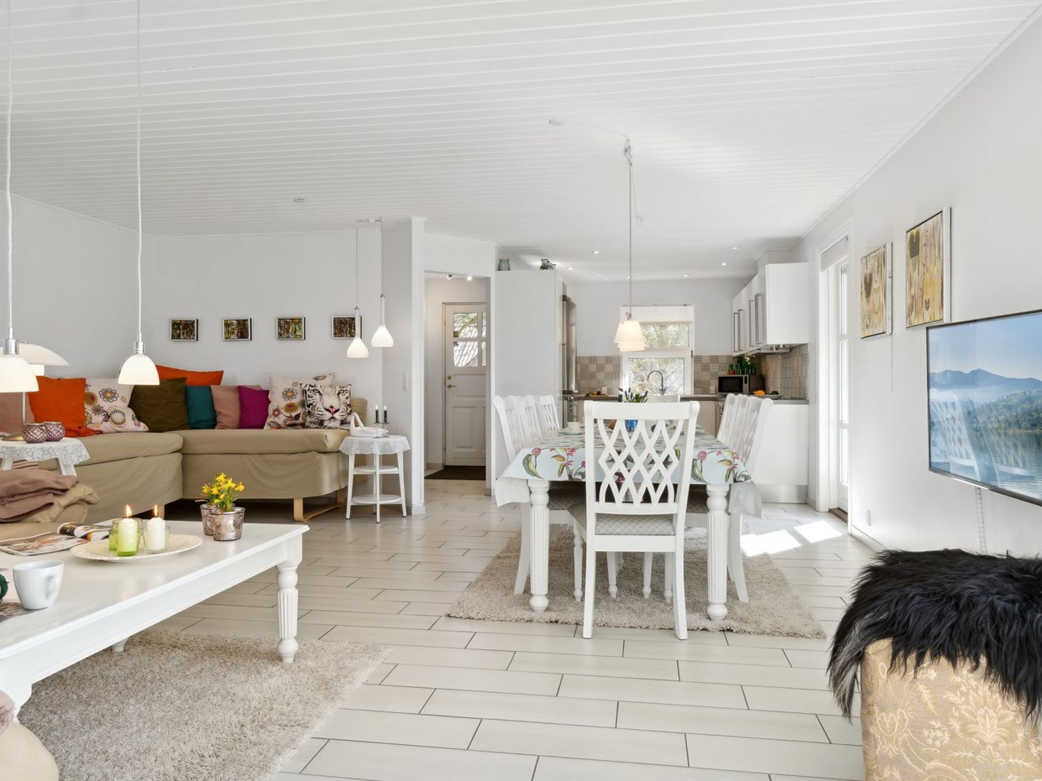 Een moderne woonkamer met een bank, eettafel en stoelen, een keuken met een televisie, en een groot raam dat zorgt voor natuurlijk licht.
