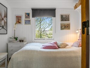 Un dormitorio con un ventanal grande, una cama con ropa de cama estampada, una mesita de noche con un florero y vasos, y decoración en la pared.