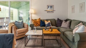 A modern living room with a green sofa, a wooden coffee table, and a white armchair. There is a glass door leading to an outdoor area.