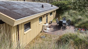 A wooden house with a sloped roof, a patio with a table and chairs, and a view of the surrounding dunes.
