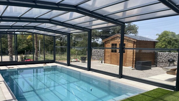 A covered swimming pool with a glass enclosure, surrounded by a stone wall and wooden structures.
