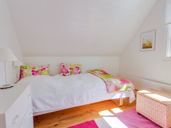 A bedroom with a bed, pillows, a bedside table, a lamp, a wicker basket, and a wall-mounted picture.