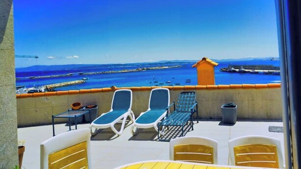 Un balcon avec deux fauteuils de jardin, une petite table et une poubelle. La vue comprend une étendue d’eau avec des bateaux et une côte lointaine.