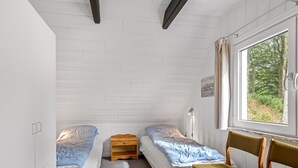 A small, single-room accommodation with two single beds, a wooden nightstand, a chair, and a window with a view of greenery.