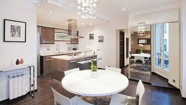 A modern kitchen with a central island, a dining area with a round table, and a view of the adjacent living space. - Magnifique 2 Pièces Parc Monceau Paris 8 (Paris)