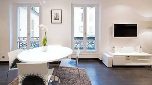 A modern living room with a round white table, two chairs, a potted plant, a flat-screen TV mounted on the wall, and a white entertainment unit. - Magnifique 2 Pièces Parc Monceau Paris 8 (Paris)