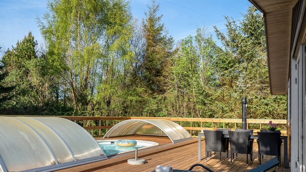 Een houten terras met een glazen hottub, lounge stoelen en een houten tafel met stoelen.