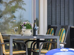 Een eettafel met een fles wijn, een glas en een bloemenvaas. Er staan rieten stoelen met gestreepte kussens.