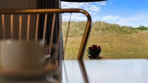 Uma mesa com xícara e pires, uma cadeira de metal e vista para um campo gramado com flores e céu claro.
