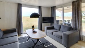 Uma sala de estar moderna com um sofá, uma mesa de centro e uma TV. Uma vista do exterior é visível através de janelas amplas.