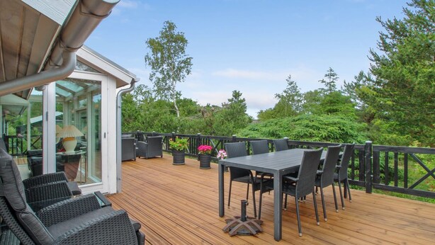 En træterrasse med udendørs møbler, en glasdør der fører til et overdækket område, og udsigt til træer og grøn vegetation.