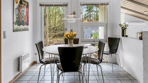 Un coin repas avec une table ronde, des chaises noires et un vase rempli de fleurs jaunes. Il y a deux fenêtres donnant sur des arbres et un mur blanc orné d’un tableau coloré.