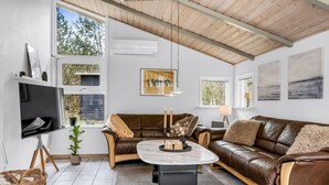 Un salon moderne avec un canapé en cuir marron, une table basse ronde et un téléviseur posé sur un meuble. De grandes fenêtres, un ventilateur de plafond et des œuvres d’art ornent les murs.
