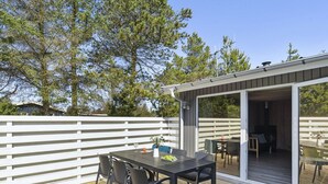 Een terras buiten met een tafel en stoelen, een overdekte zithoek en uitzicht op bomen en een gebouw op de achtergrond.