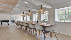 Een ruime eetruimte met een lange houten tafel en witte stoelen, grote ramen en hangende lampen.