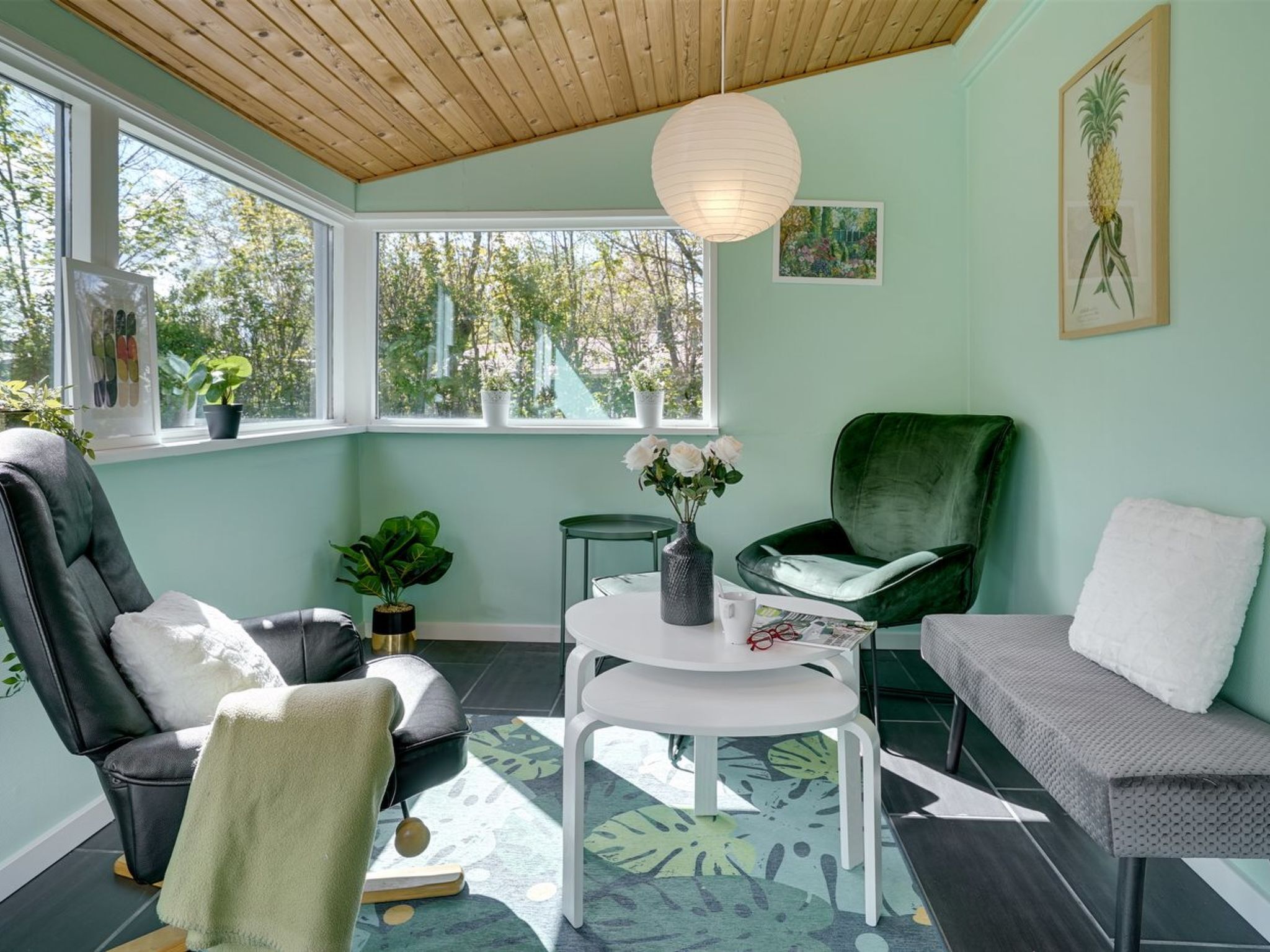 Una habitación con techo de madera, ventanas amplias, una silla verde, un sofá blanco, una mesita y una planta en maceta.
