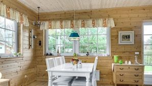 A wooden cabin interior with a dining table, chairs, a lamp, and a window with a view of greenery.