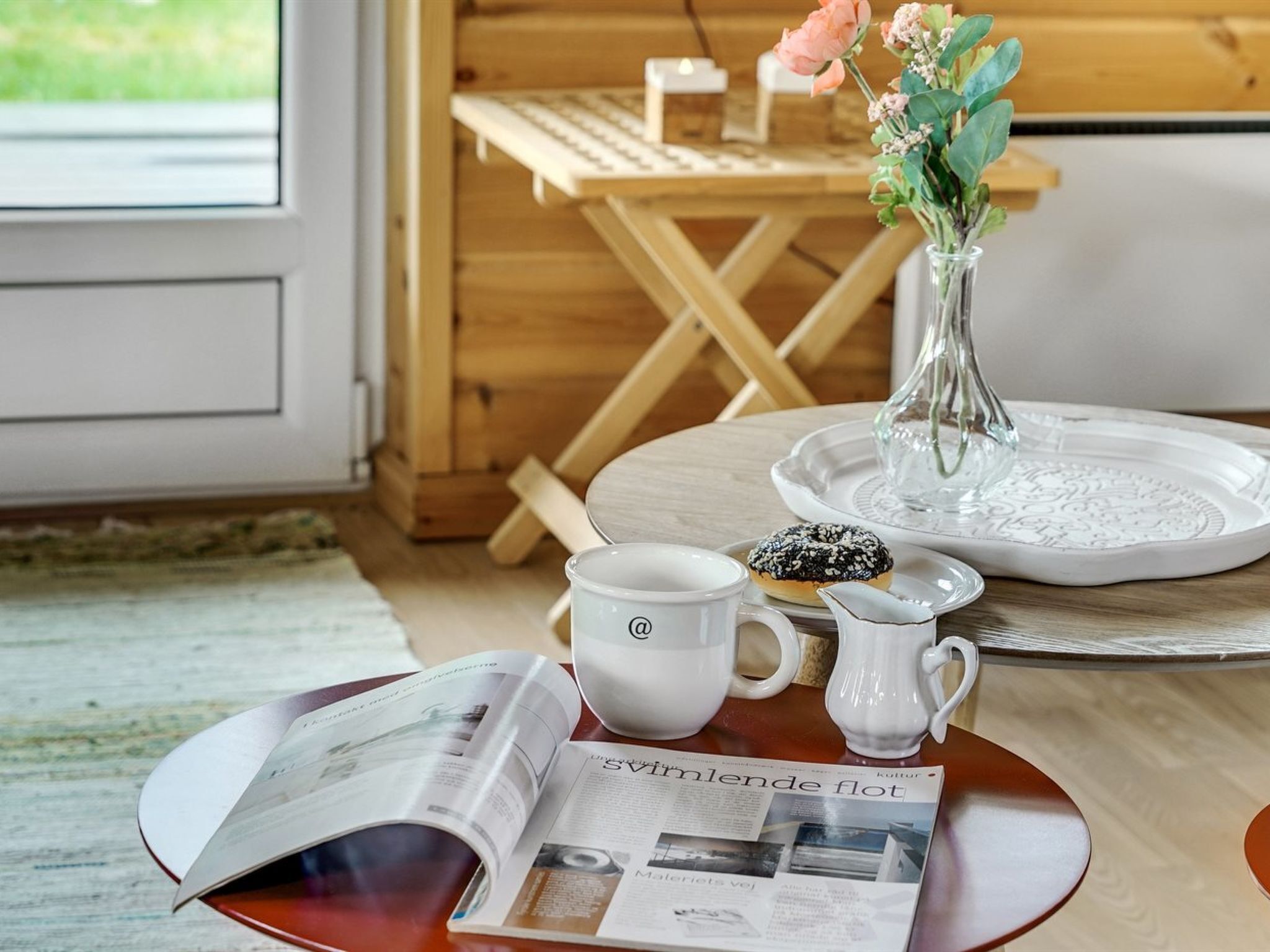 En inredning med trädetaljer och ett bord dukat för frukost, med en tidning, en mugg, en kanna och en tallrik med ett bakverk.