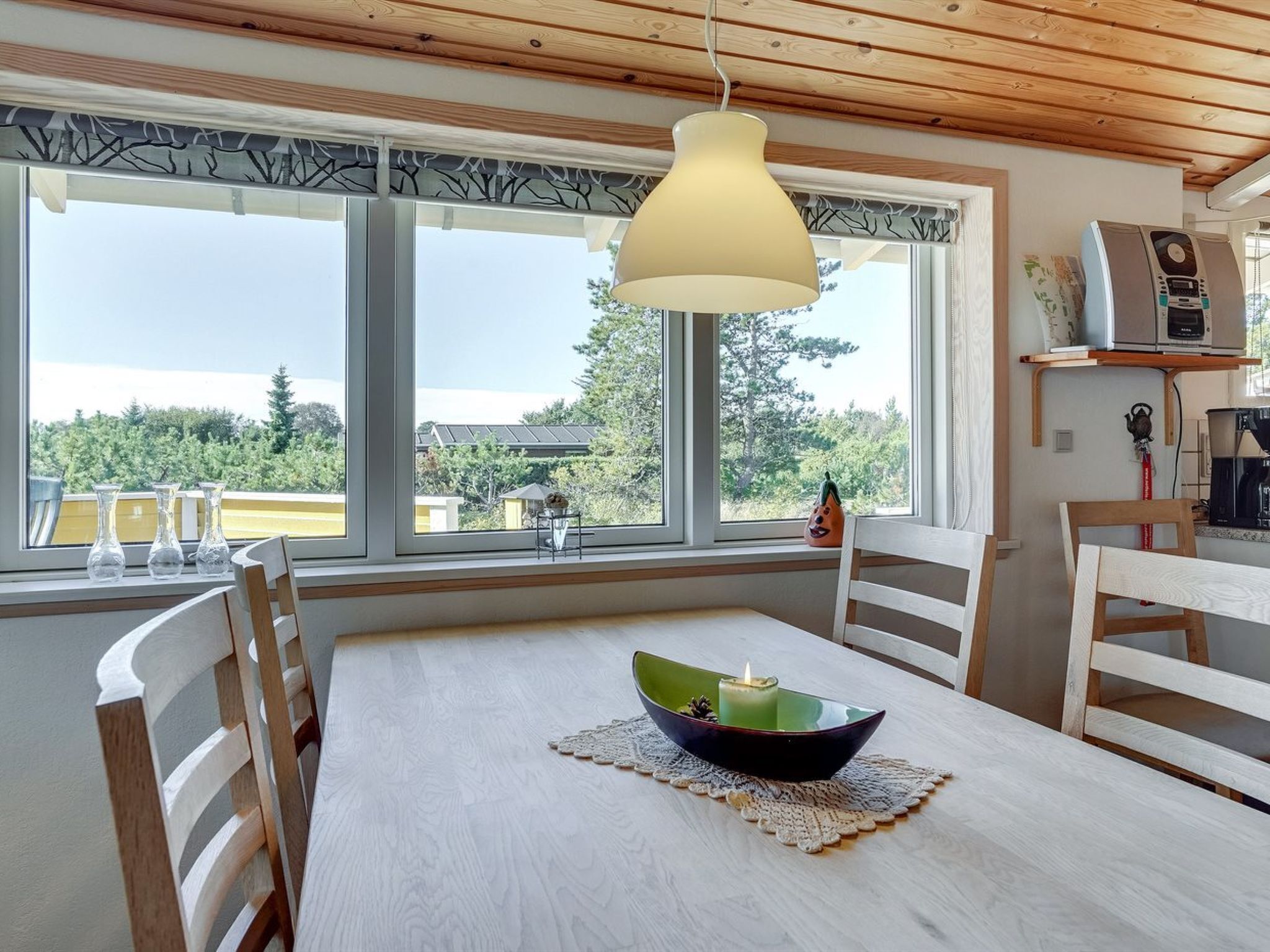 A dining area with a table, chairs, a hanging light fixture, and large windows with a view of trees and buildings.