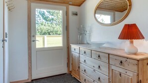 A wooden door with a view of greenery outside, a white cabinet with drawers, a round mirror, and a red lamp on a white table.
