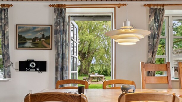 Un comedor con una mesa y sillas de madera, una lámpara colgante, un cuadro en la pared y vistas al exterior a través de amplios ventanales.