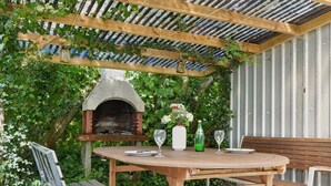 Un patio al aire libre con un horno de pizza de piedra, mesa de madera, bancos y una pérgola cubierta de follaje.