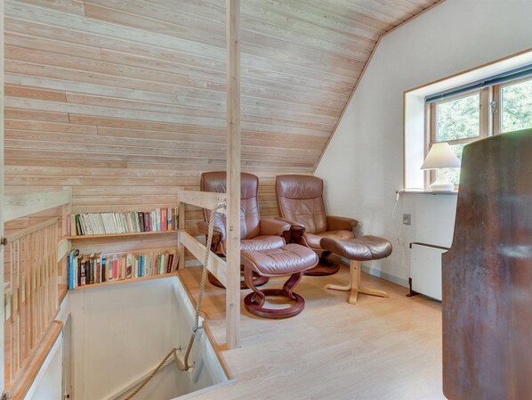 A cozy attic room with a sloped ceiling, wooden bookshelves, leather recliners, and a large window with a view of greenery.