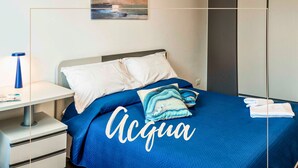 Een moderne slaapkamer met een blauwe sprei, een nachtkastje met een lamp, en uitzicht op zee vanuit het raam.